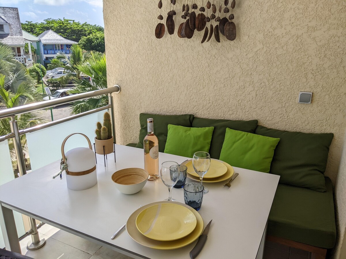 An outdoor dining area features a white table set with yellow and blue dishware, accompanied by wine glasses. Green cushions provide seating alongside the table, while a small potted cactus adds a touch of greenery. The setting is enhanced by views of the surrounding palm trees.