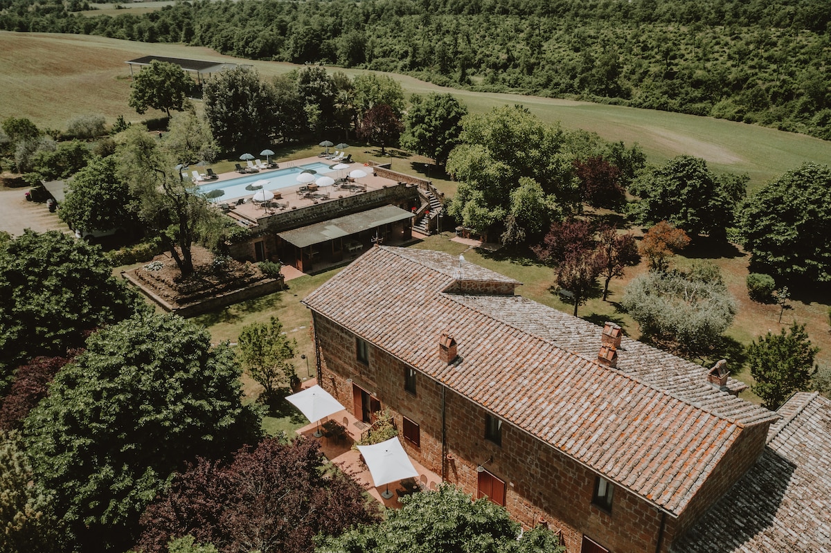 An aerial view of the Agriturismo Cerqueto showcases its rustic charm. The stone facade of the building is accented by a tiled roof. Lush greenery surrounds a shared swimming pool equipped with loungers and umbrellas, providing a tranquil outdoor space.