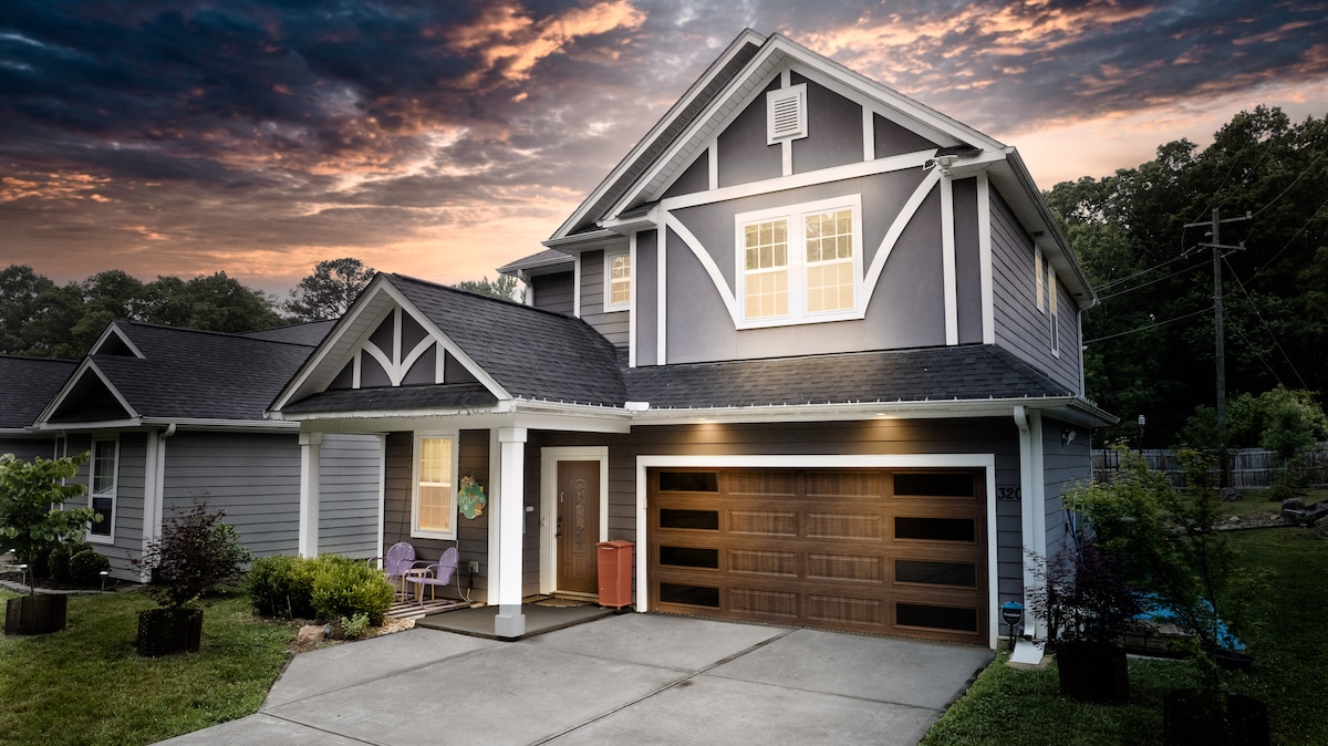 The exterior of a modern two-story home is pictured at dusk, showcasing a combination of gray siding and white accents. A welcoming front porch with two purple chairs is visible, along with a spacious driveway leading to a garage with stylish wooden doors.