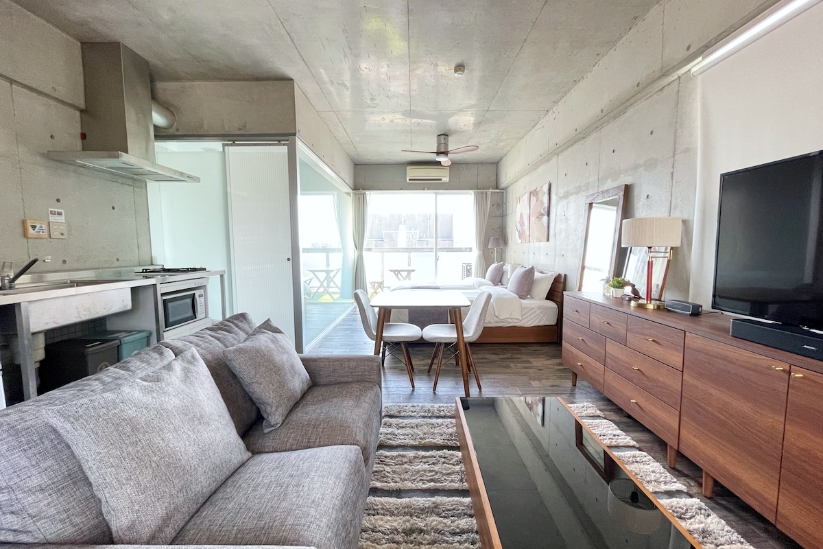An open living area is visible with a gray sofa facing a low coffee table. A dining area features a table with four chairs, and a spacious bed is positioned against a textured back wall. Natural light fills the space, complemented by sliding glass doors leading to a balcony.