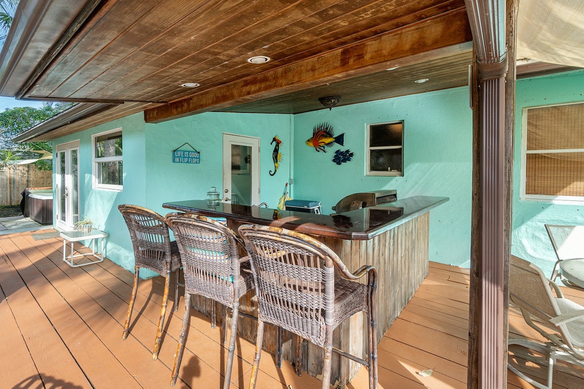 An outdoor kitchen area features a unique bar with woven stools, complemented by a vibrant aqua wall. Sunlight filters through the overhead wooden beams, creating a relaxed ambience. A view of the backyard is visible beyond, enhancing the space for entertaining.
