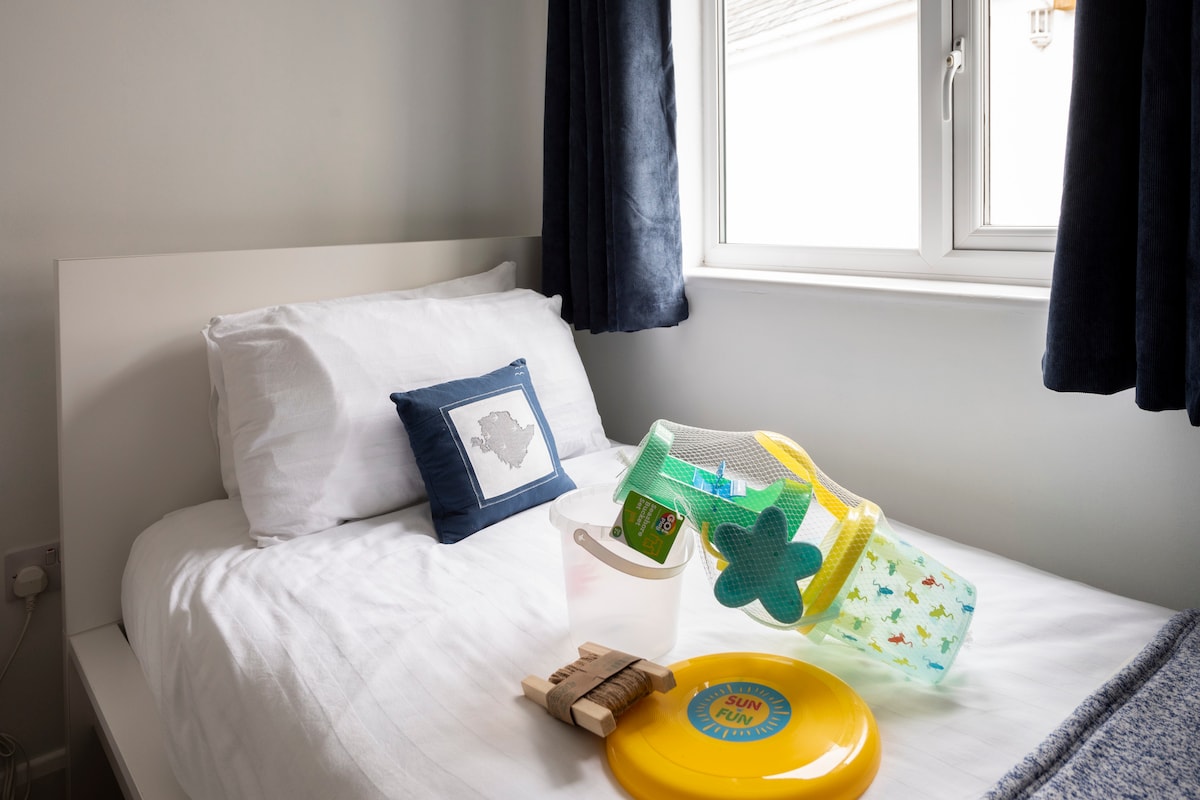 A single bed is dressed in crisp white linens, accompanied by a decorative pillow. A colorful beach toy bucket and a round frisbee are placed artistically on the bed, with sunlight filtering through a nearby window covered by dark curtains.