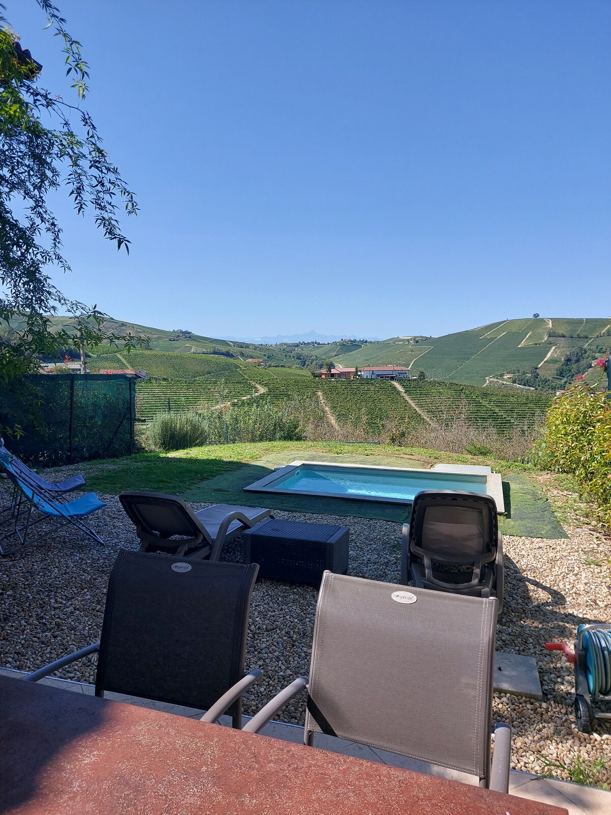 A secluded outdoor area features a small pool surrounded by a gravel patio. Two lounge chairs positioned nearby offer views of the rolling vineyards. Lush green hills rise in the background under a clear blue sky, creating a serene and private atmosphere.