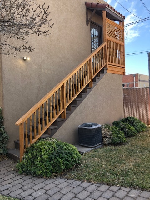 A wooden staircase with a decorative railing leads up to a door, framed by a warm-toned exterior wall. Lush green bushes and low-maintenance landscaping line the base of the steps, and an air conditioning unit is positioned at the side, offering functional cooling for the space.