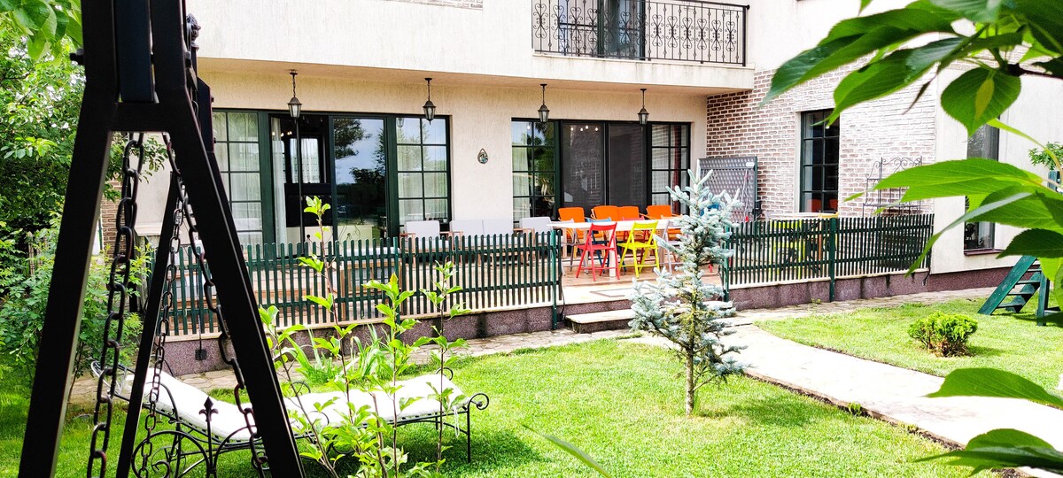 An inviting outdoor patio area features a colorful dining set surrounded by greenery. A swing set is visible in the foreground, while a well-manicured lawn leads to a pathway and the entrance of the residence, enhancing the spacious atmosphere.