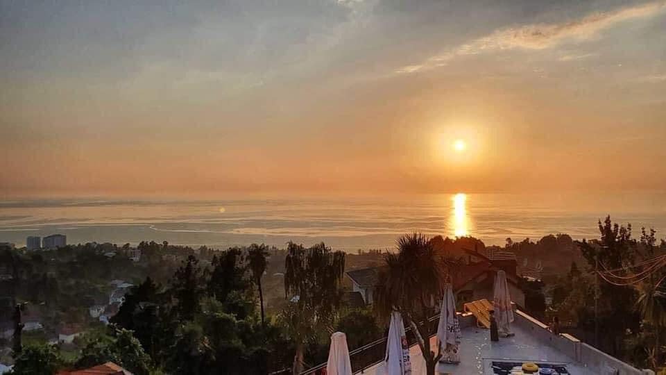 A serene sunset view is captured over the sea, with golden hues reflecting on the water's surface. Lush greenery and trees frame the scene, while distant buildings are faintly visible on the horizon. The atmosphere creates a sense of calm and tranquility.