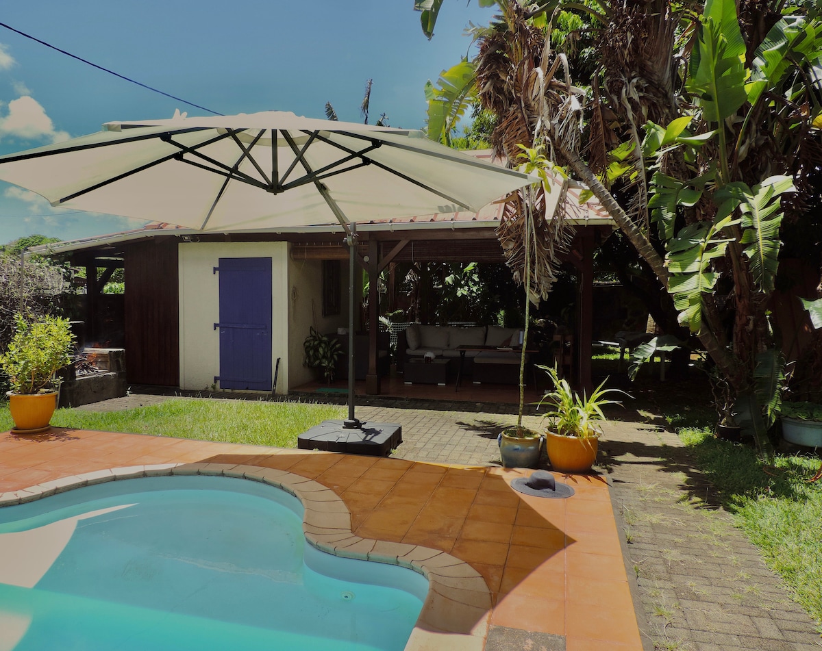 A sunlit outdoor area features a sparkling pool bordered by terracotta tiles. A large umbrella provides shade over a patio area, which is furnished with seating. Lush greenery surrounds the space, adding to the tranquil environment.