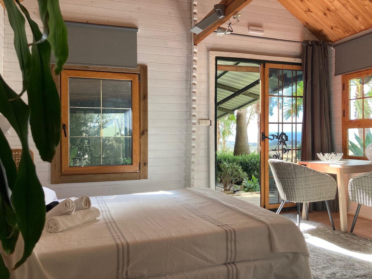 A light-filled interior features a spacious bed with neatly arranged towels. Large windows frame views of the garden, while a small dining area with two chairs and a table is visible. Natural wood beams add warmth to the decor.
