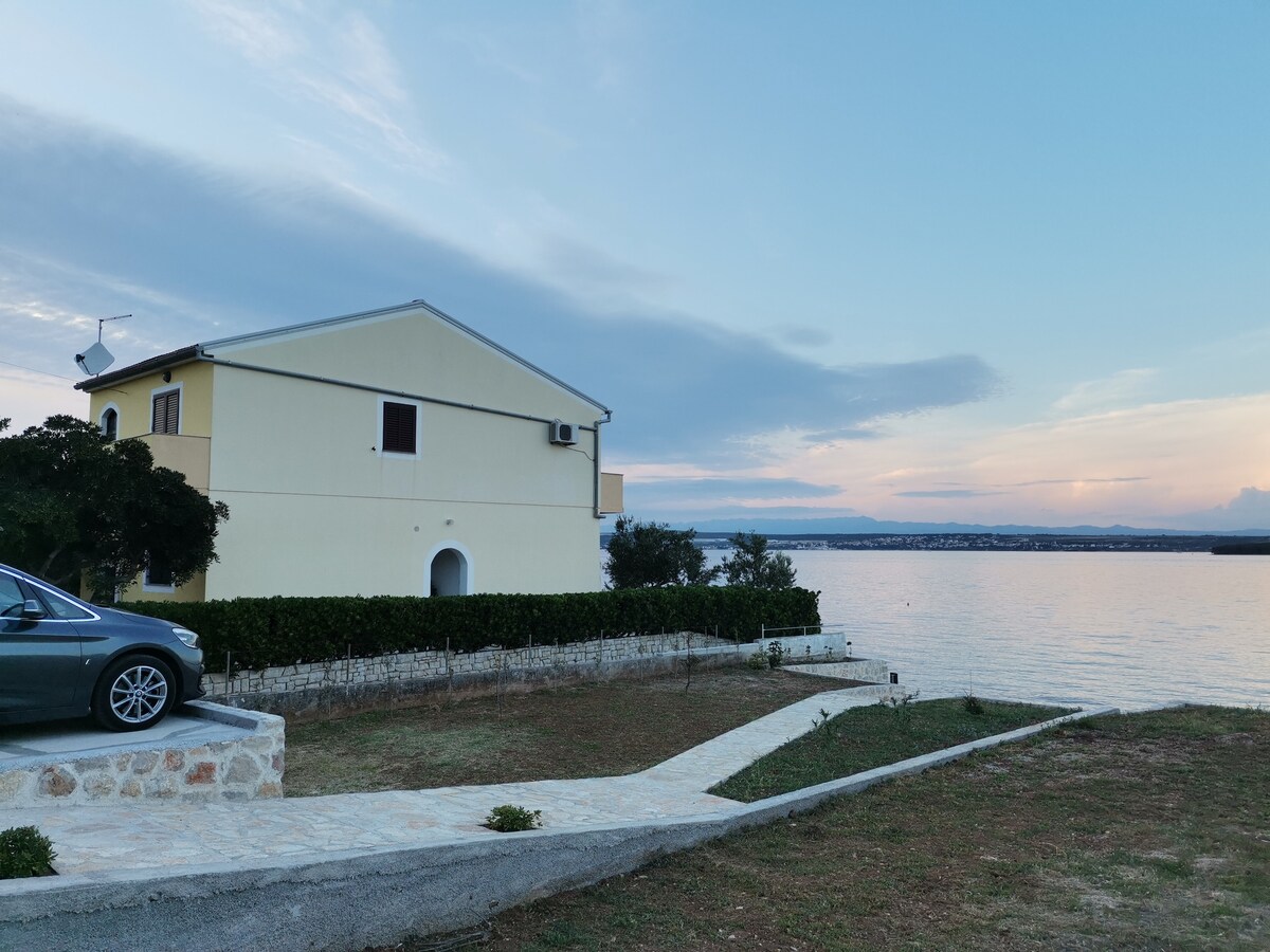 A two-story building is set beside a body of water, featuring a light-coloured exterior and multiple windows. A neatly paved pathway leads to the entrance, with surrounding greenery and a parked vehicle visible. The horizon displays soft pastel hues of sunset.
