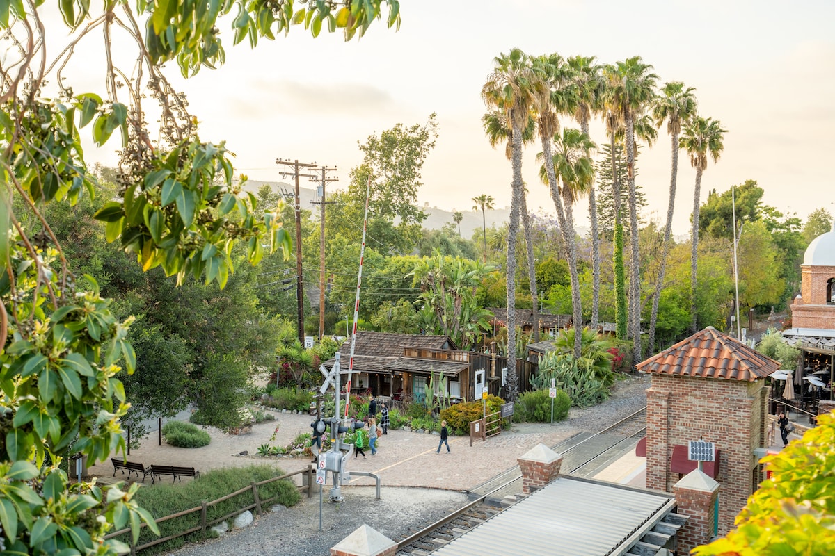The image displays a scenic view of lush greenery with palm trees surrounding rustic buildings. A pathway is visible, with people leisurely walking. The proximity to a train track is noted, enhancing the neighborhood's charming atmosphere.