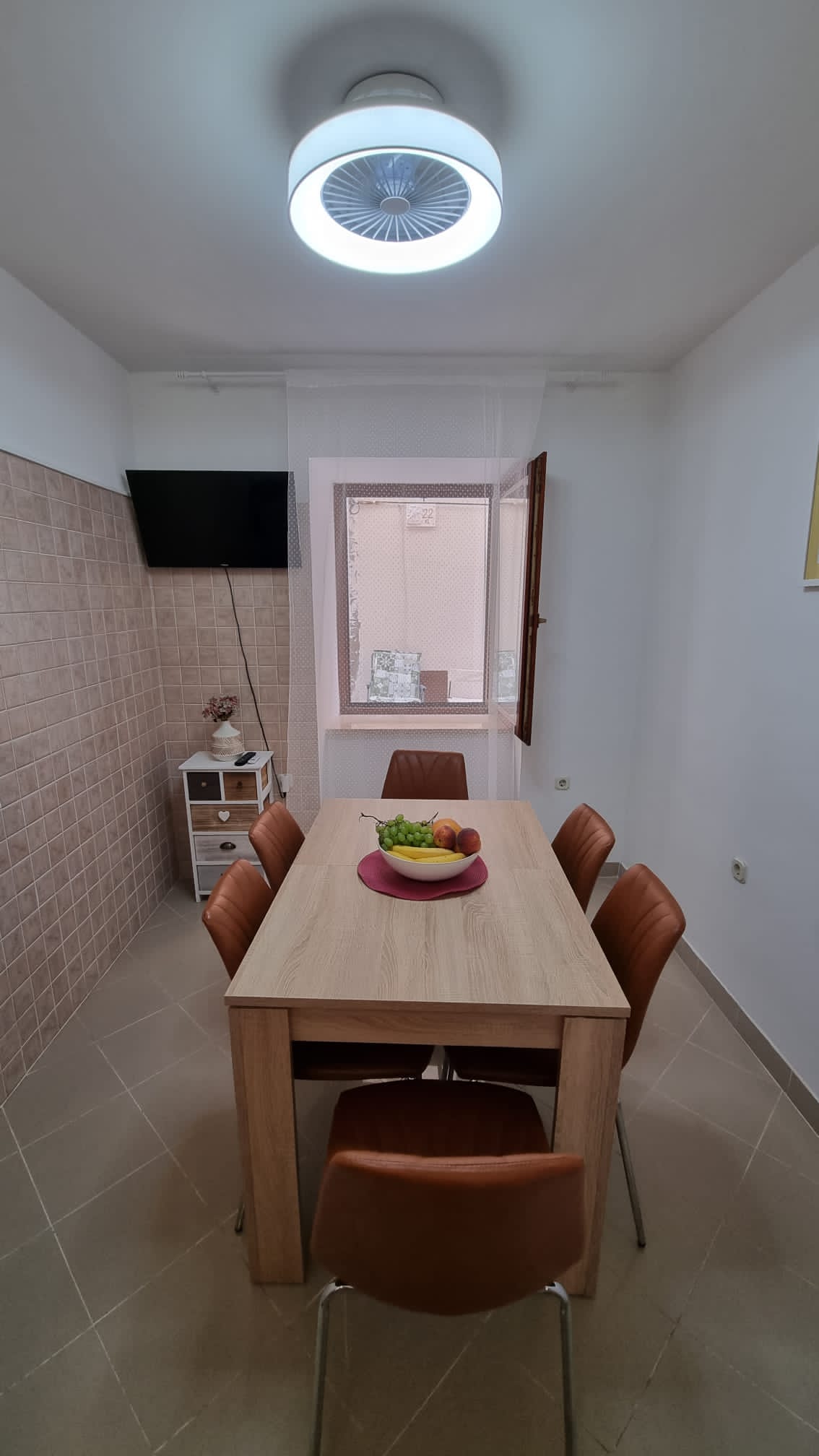 A dining area is shown with a light wooden table surrounded by six brown chairs. A bowl of fresh fruit sits at the center of the table. A wall-mounted TV is visible, and a window with pale curtains allows natural light to enter.