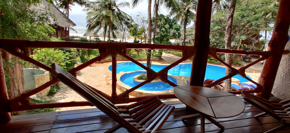 A wooden balcony provides views of a lush landscape featuring palm trees and an inviting pool. Sunbeds surround the pool area, enhancing the outdoor space toward the ocean in the distance.