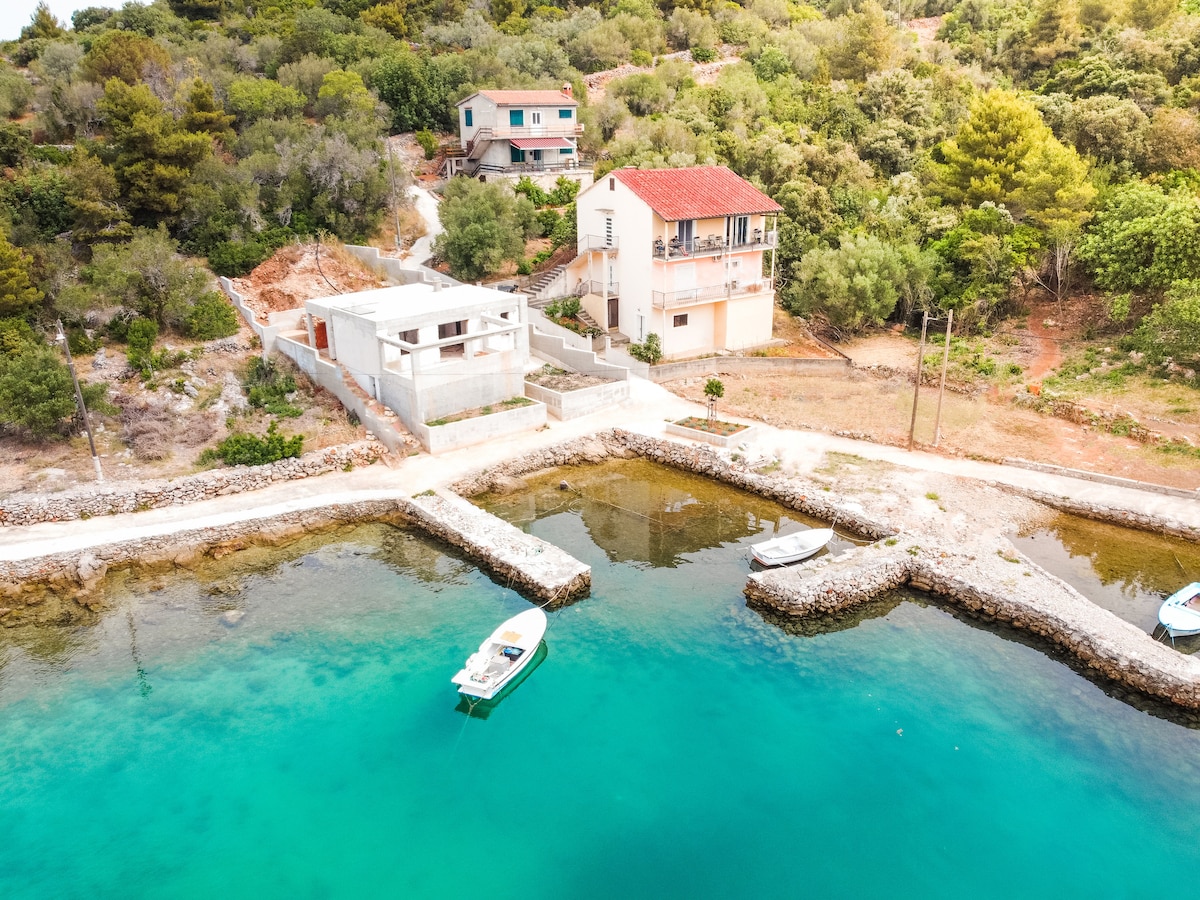 The image shows a serene seaside landscape with crystal-clear turquoise waters. A small harbor features two boats gently bobbing in the water, while a charming stone pathway leads to a house set against a backdrop of lush greenery and gently sloping terrain.