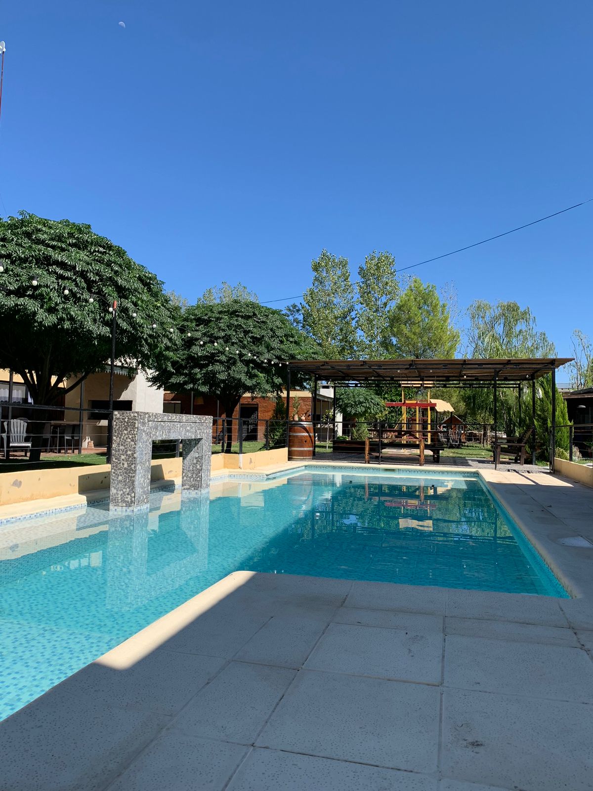 An outdoor swimming pool is surrounded by lush greenery, with shaded areas from large trees. A wooden pergola provides additional seating, creating a relaxing space by the water. The pool features a smooth stone fountain, complementing the tranquil ambiance of the area.