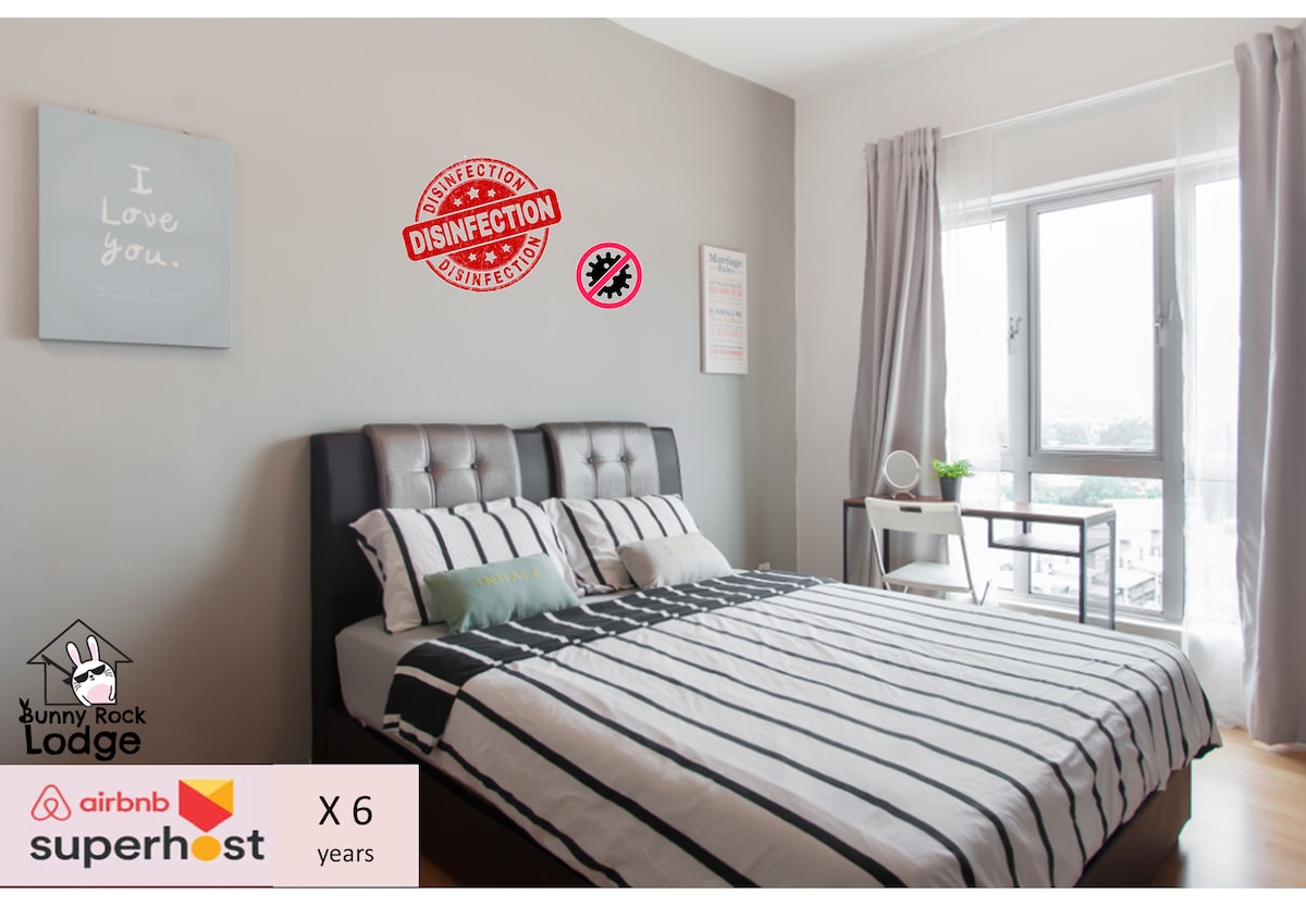 A clean and spacious bedroom features a large bed with a striped duvet and pillows. Natural light floods in through two large windows, offering views of the outside. A friendly wall decoration highlights a commitment to cleanliness and disinfection.