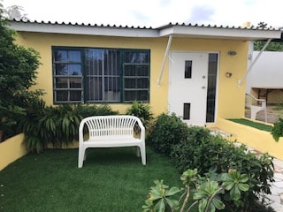 A charming yellow exterior is complemented by green framed windows and a white front door. A white bench is positioned in front, surrounded by lush greenery and well-maintained landscaping. A small patch of artificial grass enhances the inviting entrance.