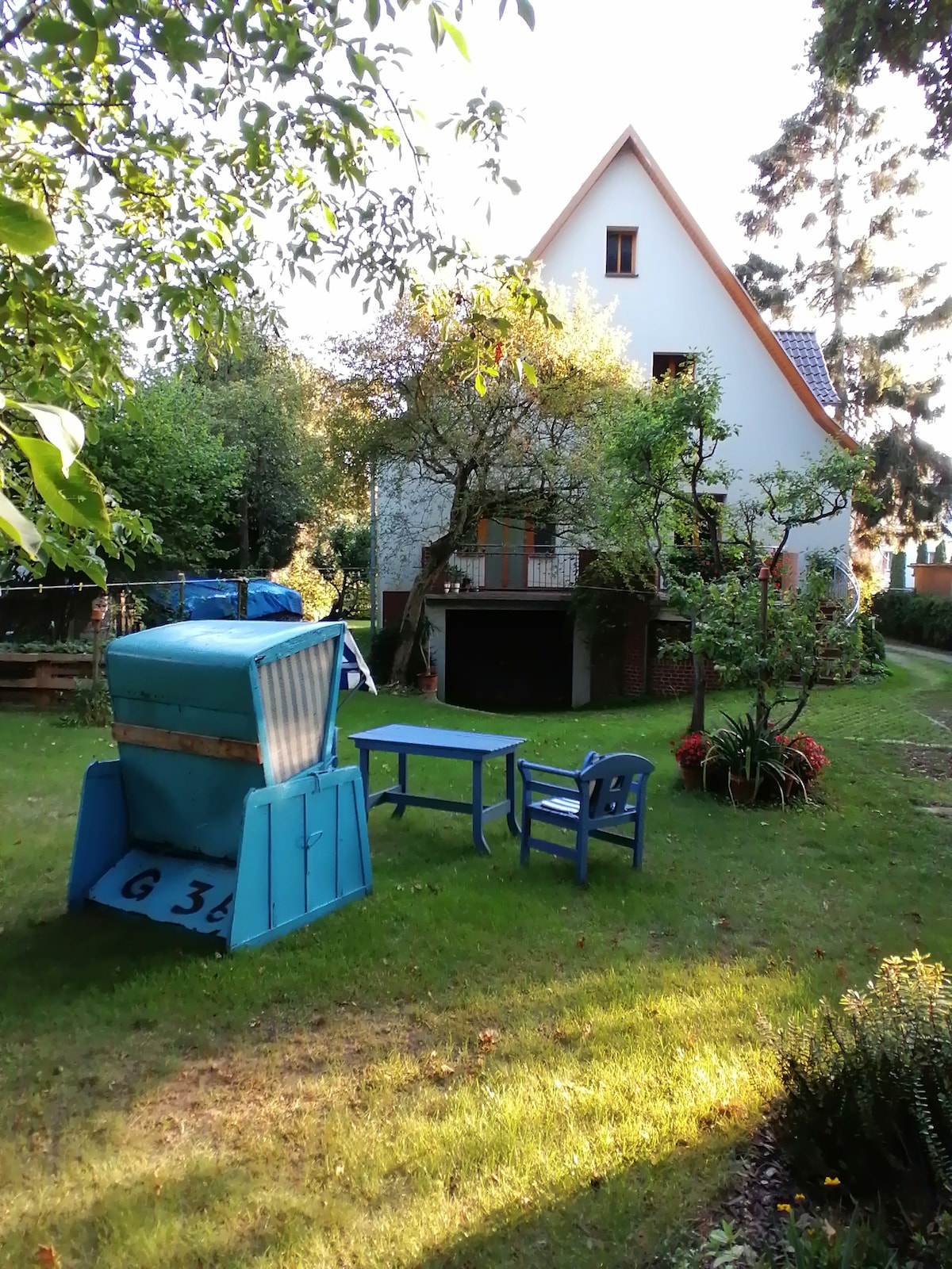 A large garden surrounds the house, featuring a blue storage unit and a small table with two chairs. Lush green grass and colorful flowers are present, creating a serene outdoor space. The house, characterized by a sloped roof and large windows, is partially shaded by trees.