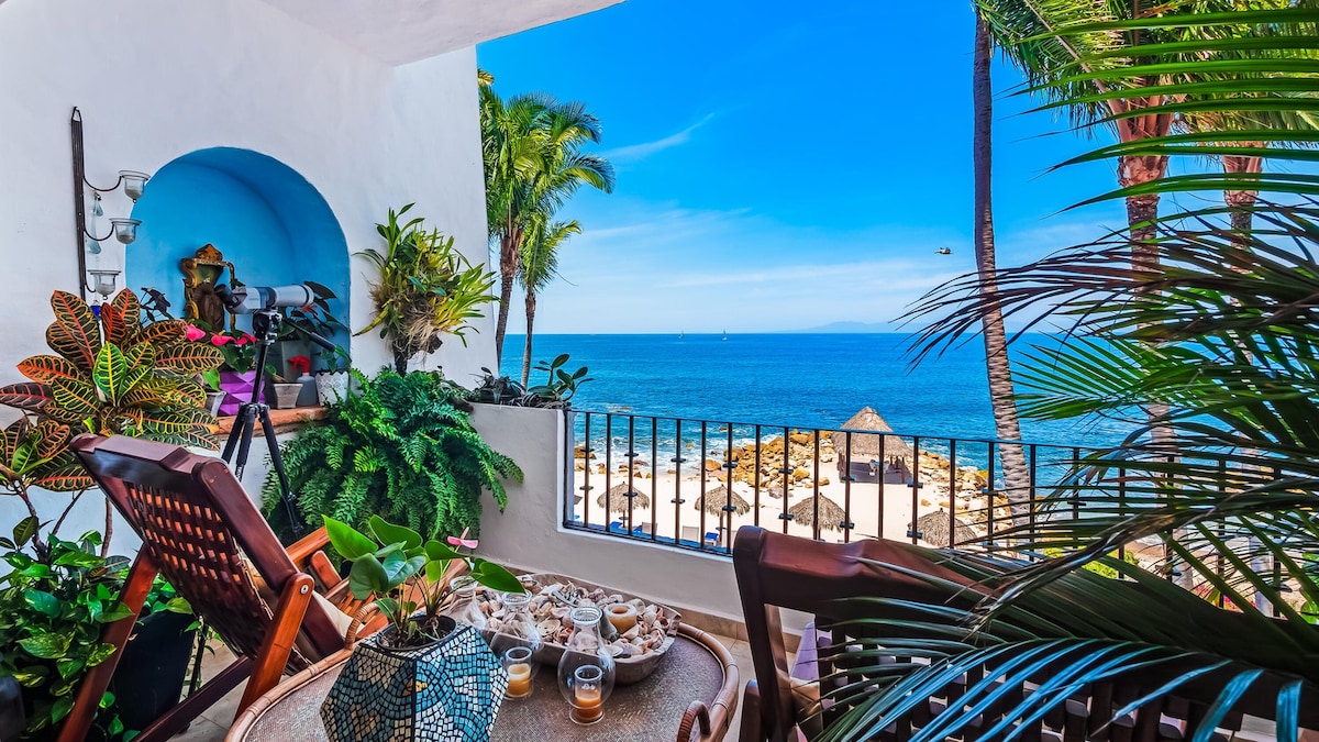 A serene balcony offers views of the ocean, framed by vibrant palm trees and lush greenery. A round table is set with plates and drinks, surrounded by comfortable seating. The distant beach features umbrellas, enhancing the coastal atmosphere.