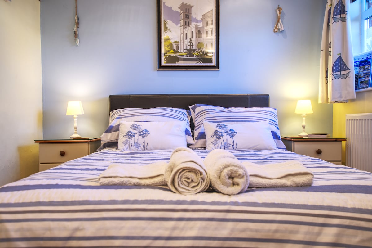 A cozy bed is dressed in striped linens, flanked by two bedside lamps providing soft illumination. Fresh towels are neatly arranged at the foot of the bed. A framed artwork featuring coastal scenery hangs above, lending a serene touch to the room.