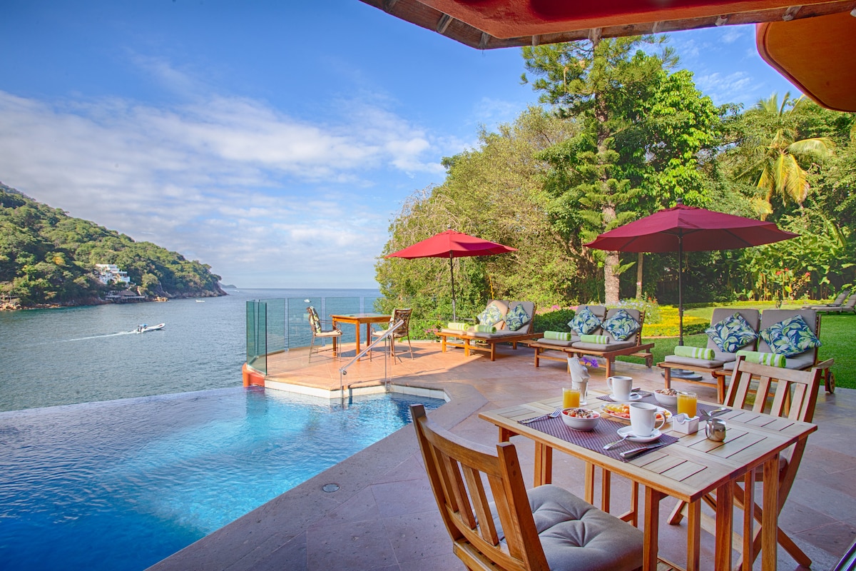 An inviting outdoor space features a tranquil infinity pool, surrounded by lounge chairs and parasols. The nearby shoreline is visible, along with lush greenery that enhances the serene environment. A table is set for breakfast, creating a perfect spot to enjoy the view.
