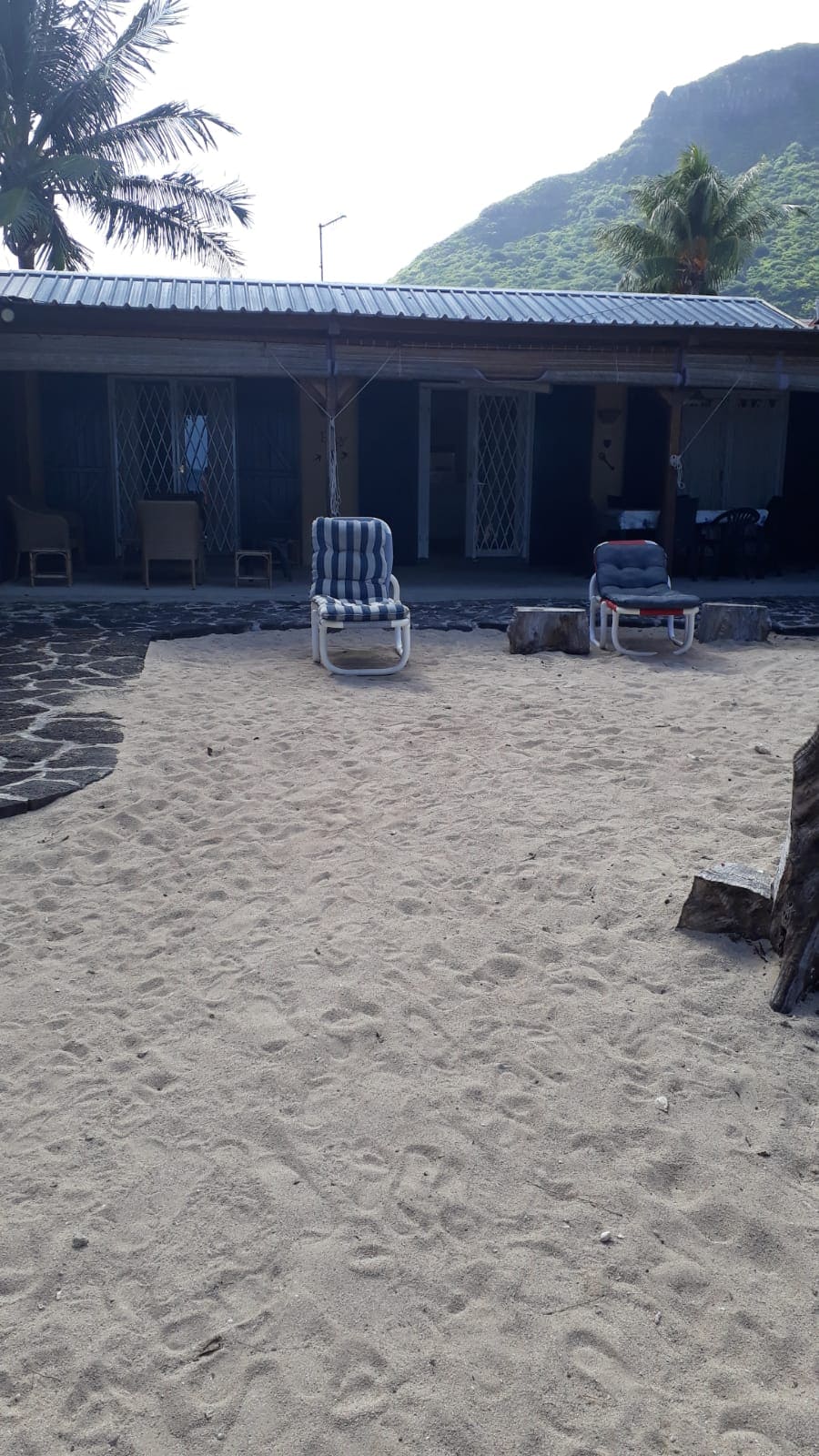 A sandy outdoor area is presented in front of the cottage, featuring two lounge chairs positioned for relaxation. The entrance of the cottage is visible, with a shaded porch area providing access. Natural stone elements frame the scene, enhancing the beachside environment.
