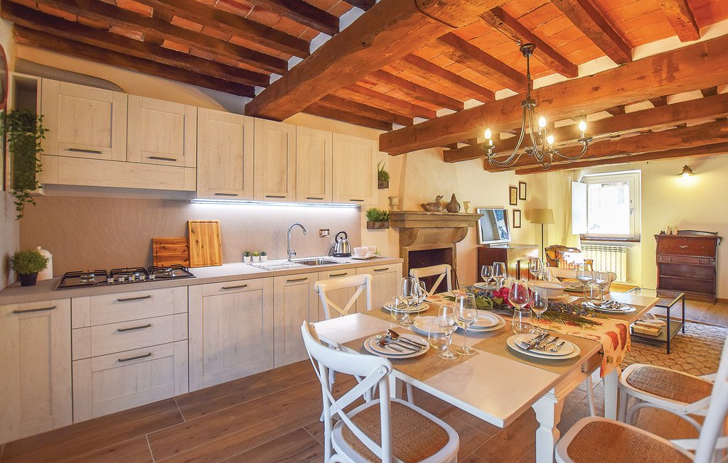 A spacious kitchen area features wooden beams overhead and light-colored cabinetry. A dining table is set with plates and glassware, accompanied by a cozy seating arrangement. A large window allows natural light to fill the room, creating a warm and inviting atmosphere.