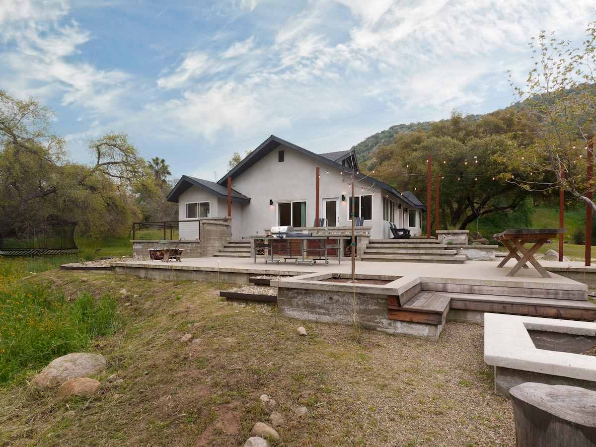 An outdoor patio area features a spacious deck with seating arrangements, surrounded by greenery and elegant string lights. A fire pit and wooden picnic table are included, creating a relaxed atmosphere for gathering. The backdrop consists of rolling hills and trees.