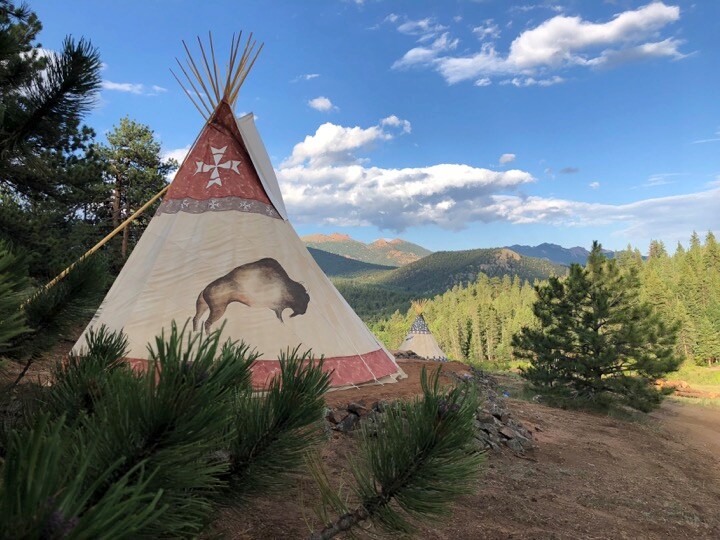 Two tipis are set amidst a vast landscape of trees and mountains. One tipi features a bison design, while the other stands in the background. The sky is partly cloudy, and pine branches frame the scene, enhancing the natural surroundings.
