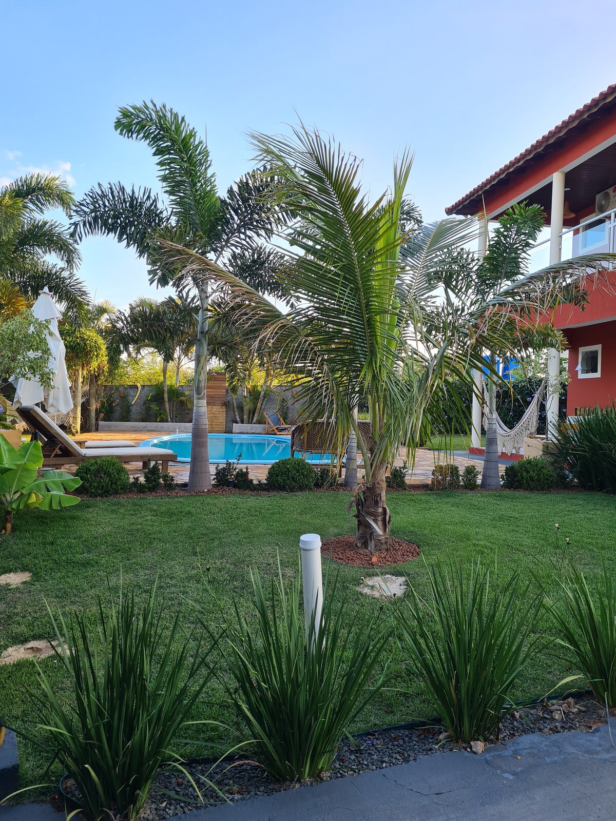 A vibrant garden area showcases well-maintained green grass and tropical plants, including palm trees. A swimming pool is visible in the background, surrounded by wooden decking and lounge chairs. The exterior of the house and a clear blue sky complement the serene outdoor setting.