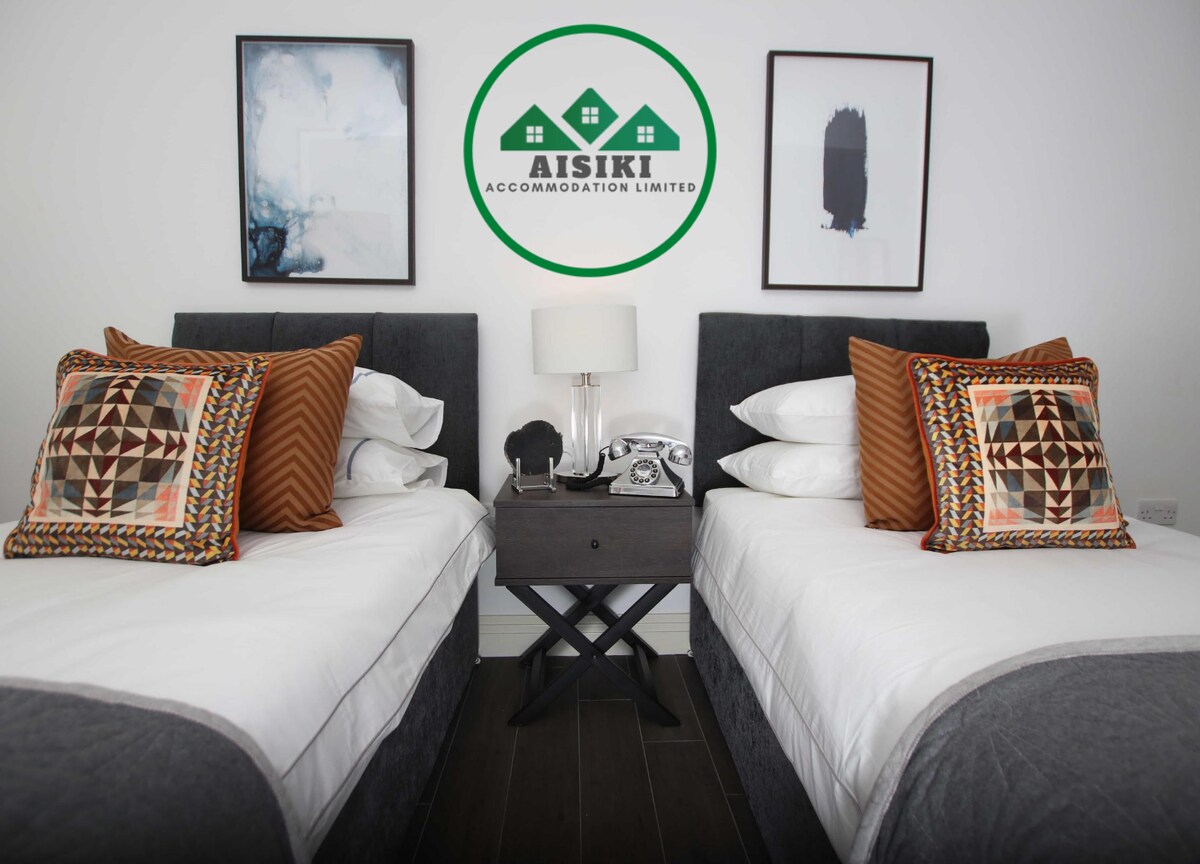 The image shows a well-appointed bedroom featuring two spacious twin beds, each adorned with patterned cushions and crisp white linens. A bedside table with a lamp and a vintage telephone is positioned between the beds, against a backdrop of framed art on the walls.