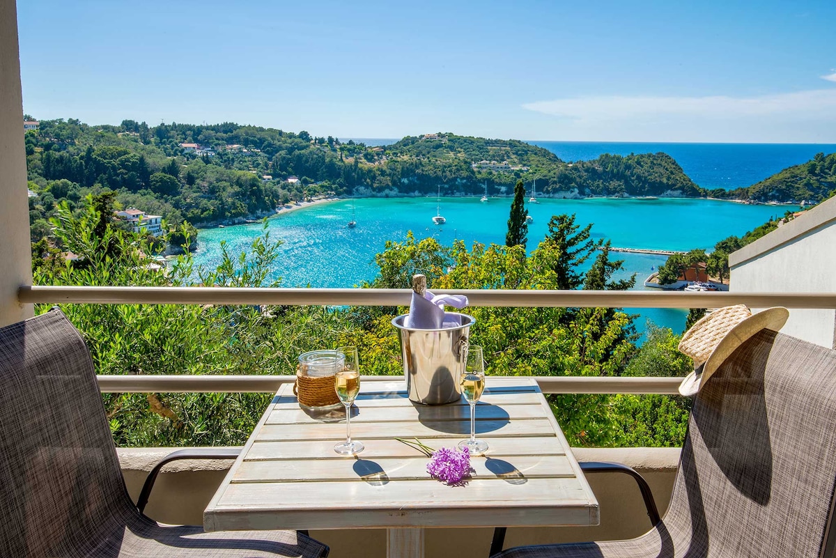 A terrace table is set for two, featuring a bucket of chilled drinks and glasses. Chairs face stunning views of Lakka Bay, framed by lush greenery. The vibrant blue waters and distant hills provide a serene backdrop, enhancing the calm atmosphere.