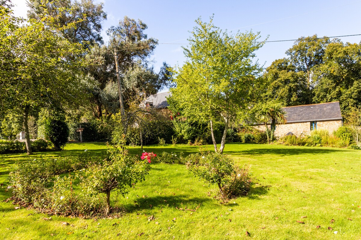 A spacious garden is surrounded by trees and shrubbery, featuring a well-maintained lawn. Flower beds with blooming plants are visible, and a stone barn can be seen in the background, blending harmoniously with the natural setting.