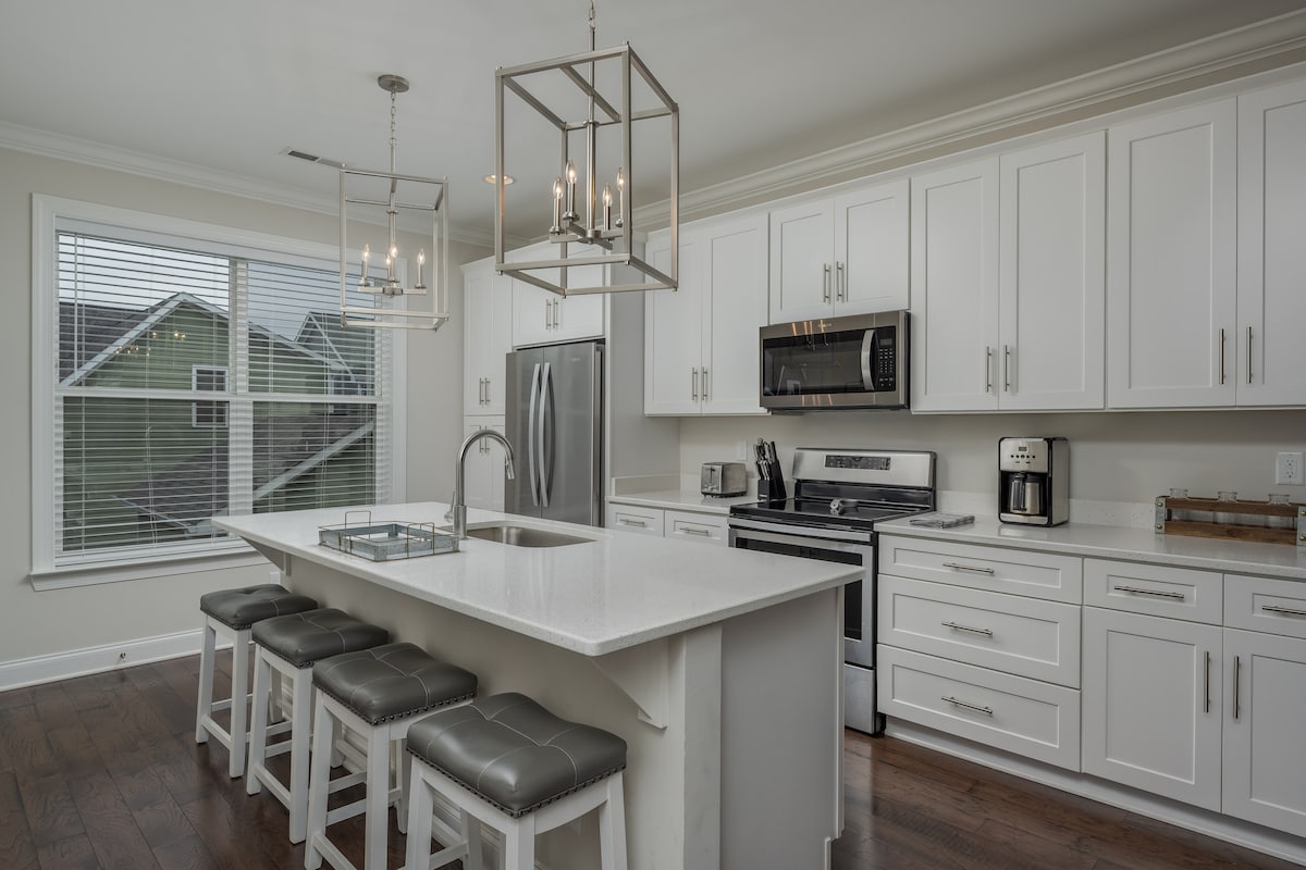 A modern kitchen features white cabinetry and stainless steel appliances, including a refrigerator and microwave. An island with seating for four is complemented by stylish pendant lights. Nearby, a window offers natural light and a view of the outdoors.