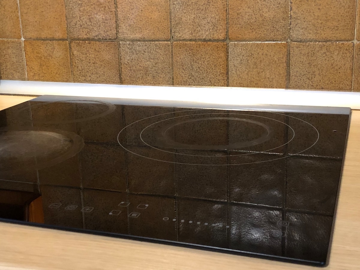 A smooth cooktop is displayed, featuring circular heating elements against a reflective black surface. The backsplash behind the cooktop consists of textured brown tiles, providing a warm contrast to the sleek design.