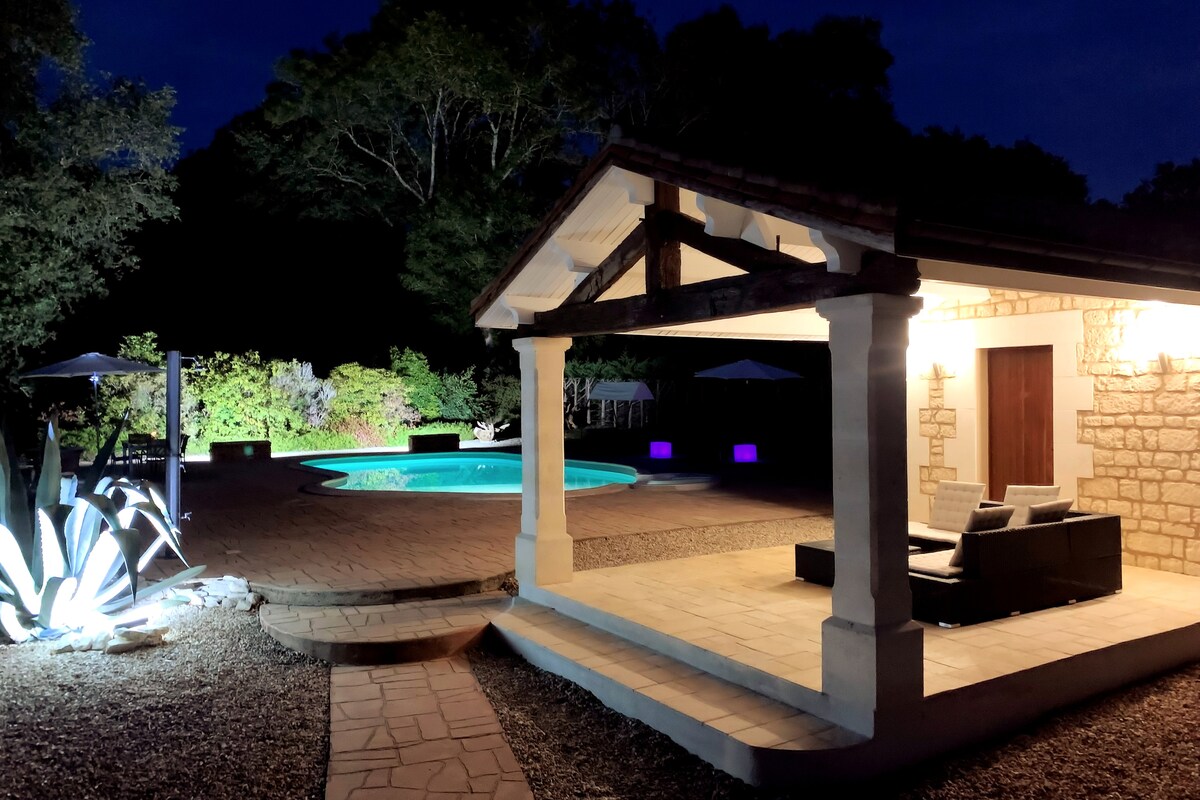 An outdoor area features a poolside view with a well-lit pool set against dark foliage. A covered seating area is visible, with furnishings arranged for relaxation. Soft lighting highlights the structure, creating a serene atmosphere for evening enjoyment.