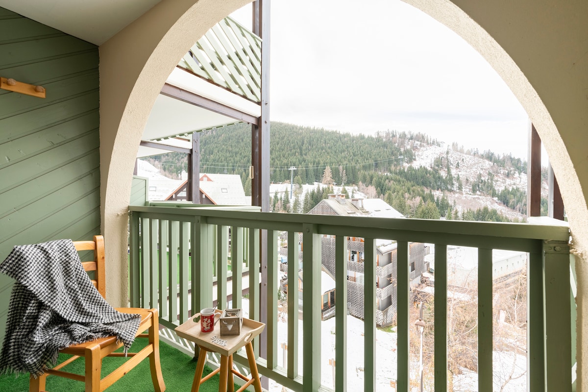A balcony is framed by an arched opening, featuring a wooden chair and a small table. A cozy blanket rests on the chair. Scenic views of snow-covered hills and nearby buildings are visible in the background, providing a serene outdoor space.