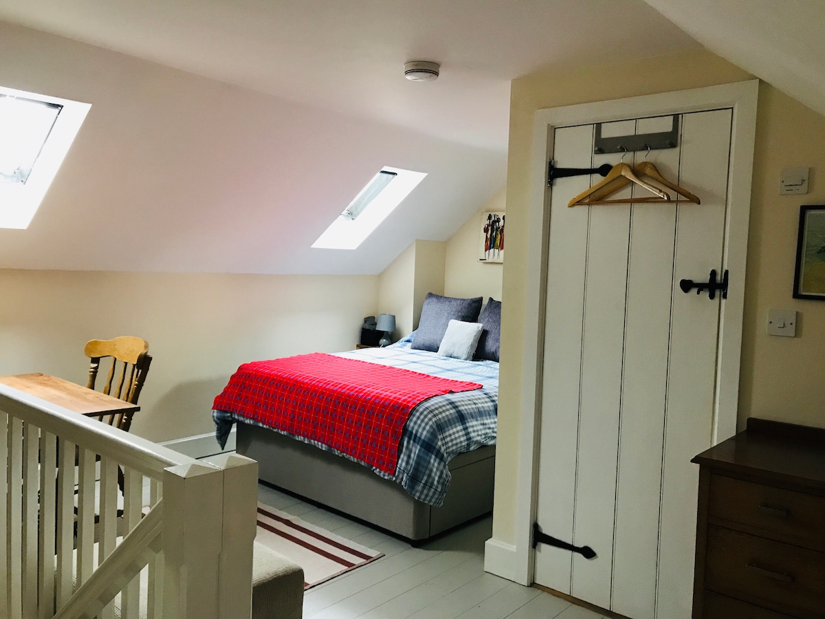 A cozy attic room features a spacious bed dressed in a plaid blanket. Natural light filters through two skylights, illuminating a small wooden table and chair set. A white door with rustic hardware adds a charming touch to the neutral decor.