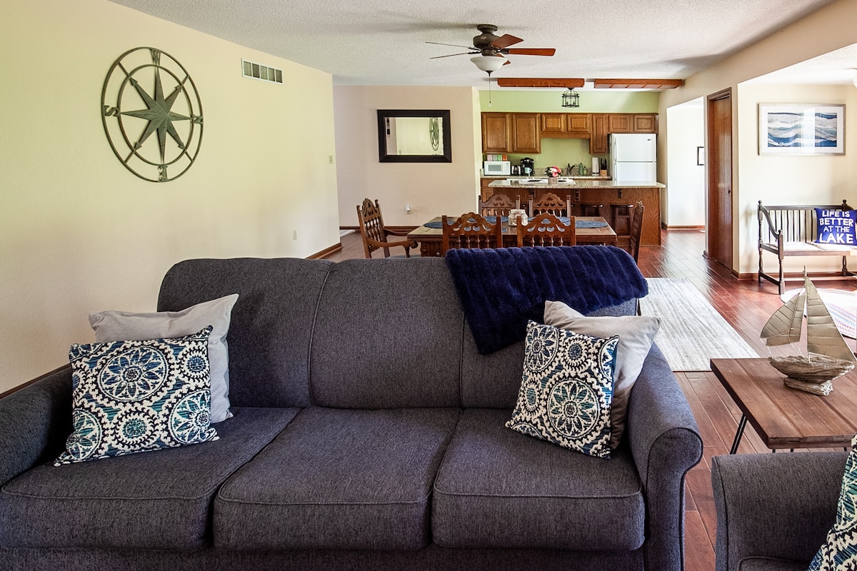 A spacious living area displays a comfortable gray sofa adorned with decorative pillows. A dining table is visible in the background, paired with wooden chairs. A kitchen with wooden cabinetry and a white refrigerator is located beyond. Natural light streams through the windows.