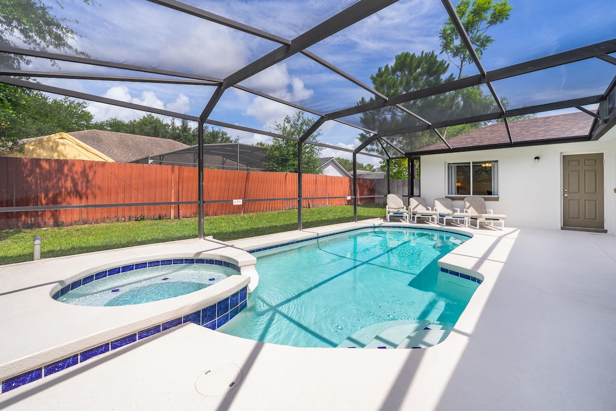 The private pool area is fully screened in, featuring a shimmering pool and a separate spa. Sun loungers are positioned by the water, surrounded by a spacious deck. Tall hedges and a fence create a sense of privacy, complemented by clear blue skies.