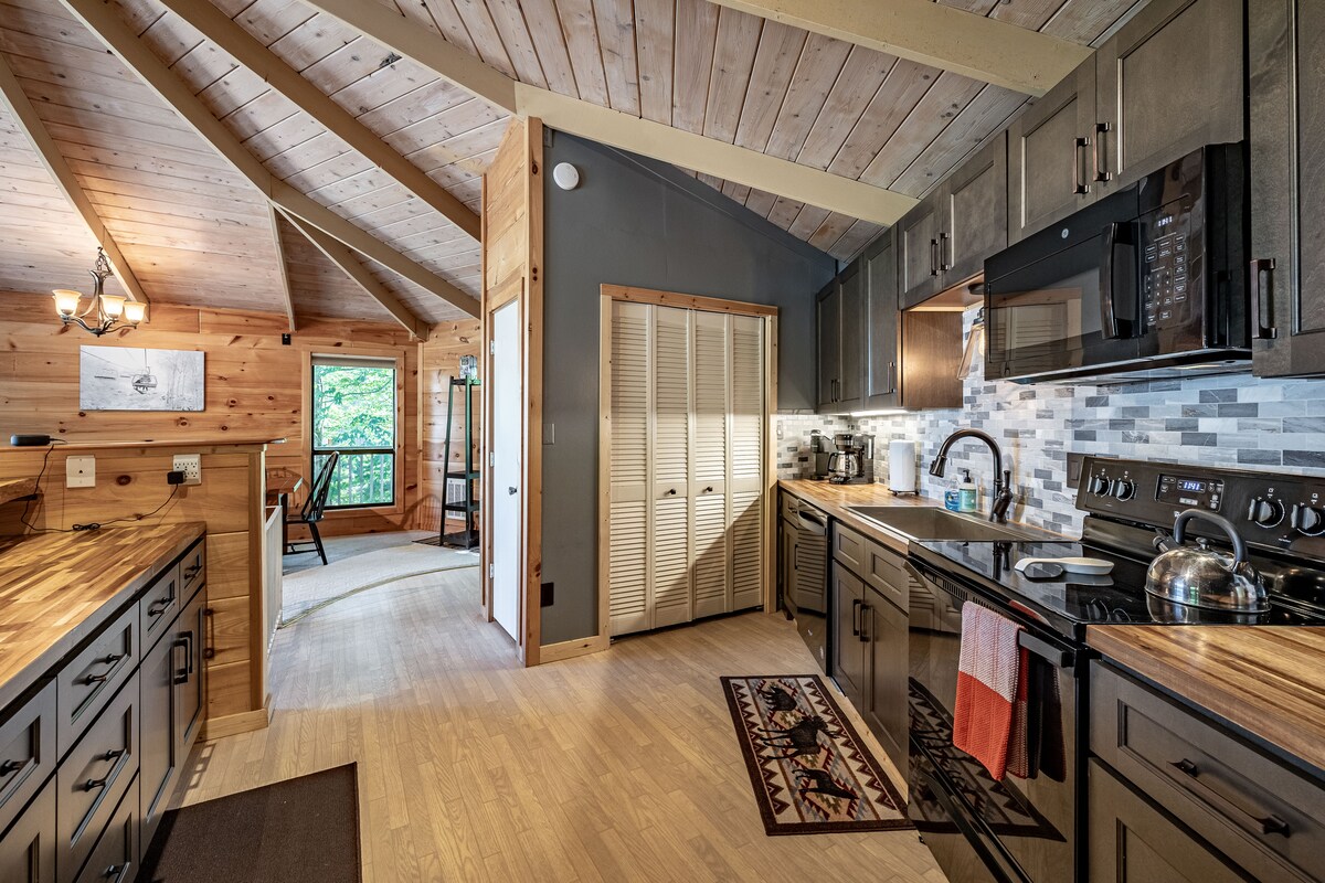 A spacious kitchen features wooden cabinetry and sleek black appliances. Ample counter space is complemented by a decorative area rug on the floor. Large windows allow natural light to fill the space, while wood-beamed ceilings add warmth to the inviting atmosphere.