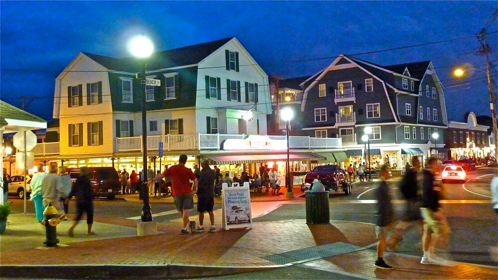 A bustling street scene is captured at night, featuring charming, multi-story buildings with large windows. People are visible walking along the sidewalk, while streetlights cast a warm glow. A nearby restaurant's bright signage stands out, adding to the lively atmosphere.