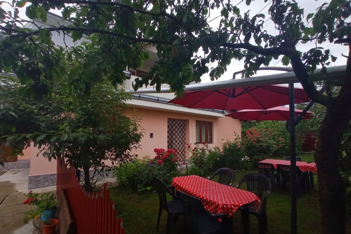 A charming outdoor area features several tables covered with red tablecloths, situated beneath large umbrellas. Lush greenery and flowering shrubs surround the space, contributing to a relaxed atmosphere. The exterior of the house is painted in soft pink, with windows framed in white.