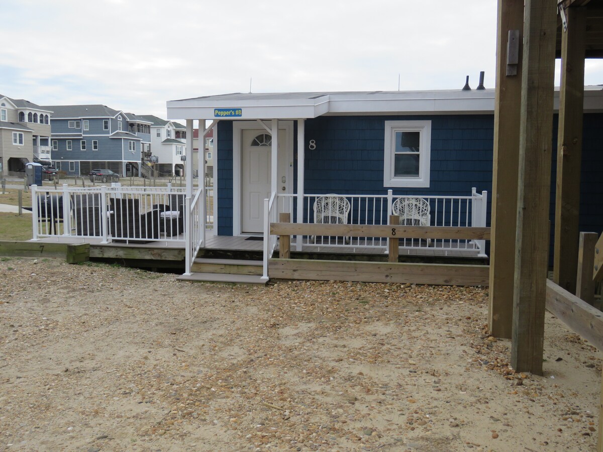 An exterior view of Pepper's Cottage showcases a blue wooden structure with a white railing and entryway. Two white chairs are positioned on the porch, offering a space to relax. The sandy area in front leads to neighboring cottages, enhancing the charming coastal atmosphere.