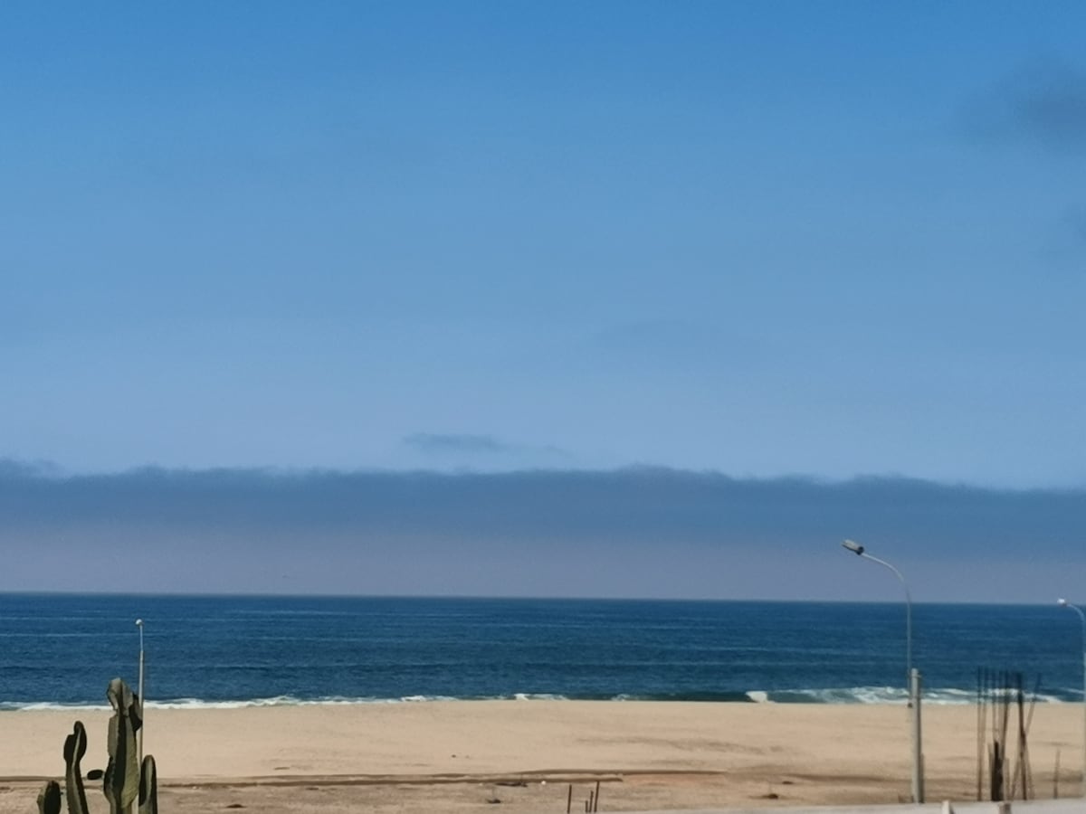 The image captures a wide view of a sandy beach meeting the ocean under a clear blue sky. Gentle waves are visible along the shoreline, with distant clouds softly blending into the horizon. A few lampposts stand along the beach, adding to the coastal scenery.