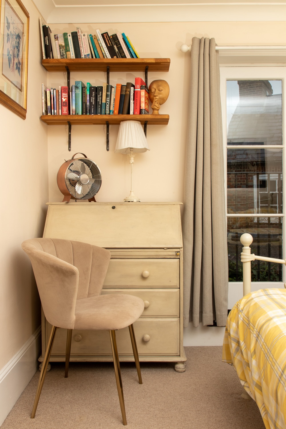 The room features a quaint writing desk with drawers and a light-colored finish. Above, a shelf holds a collection of books, accompanied by a small lamp and a decorative fan. A cozy chair with a soft texture is positioned nearby, complementing the inviting atmosphere.