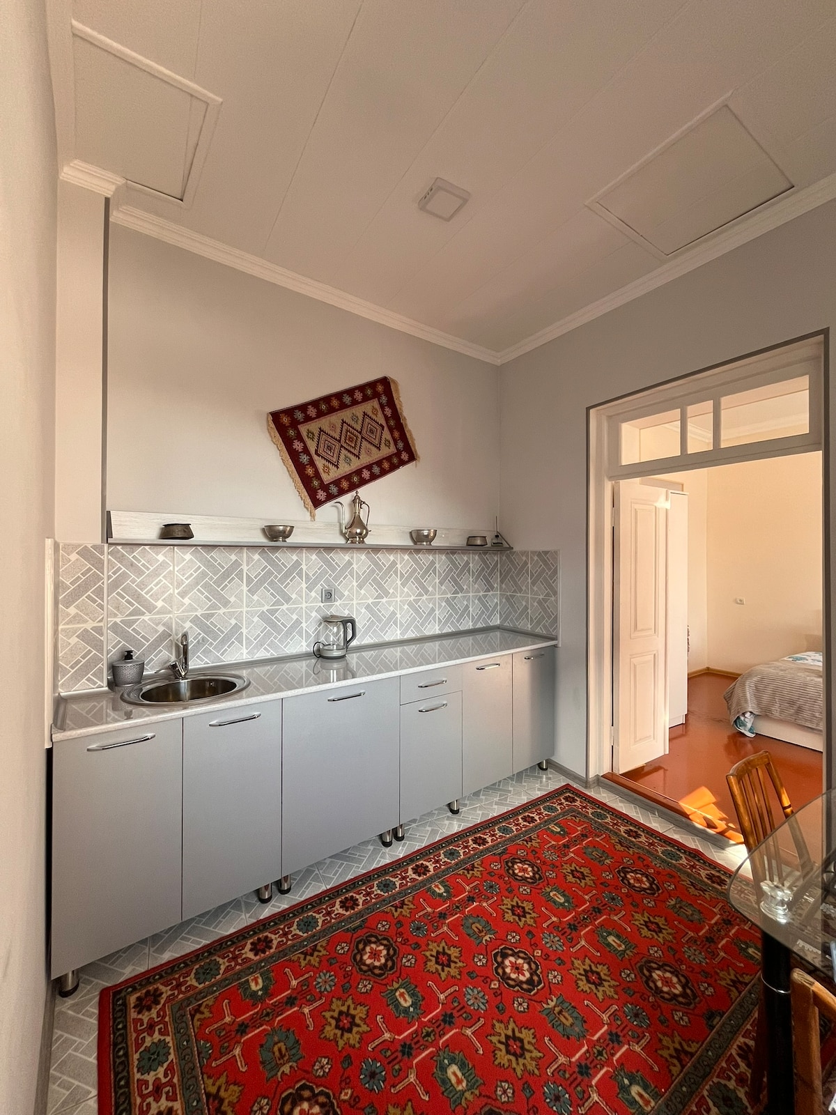 A compact kitchen features modern cabinetry in a soft gray finish, complemented by patterned tiles. A colorful oriental rug lies on the floor, adding warmth to the space. An entrance to a separate room is visible through an open door.
