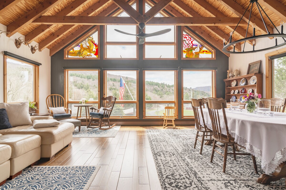 A bright great room features vaulted wooden ceilings and large windows framing mountain views. A cozy sectional sofa is placed beside a patterned area rug. A dining table with wooden chairs occupies one area, while a small table with rocking chairs is positioned near the windows.