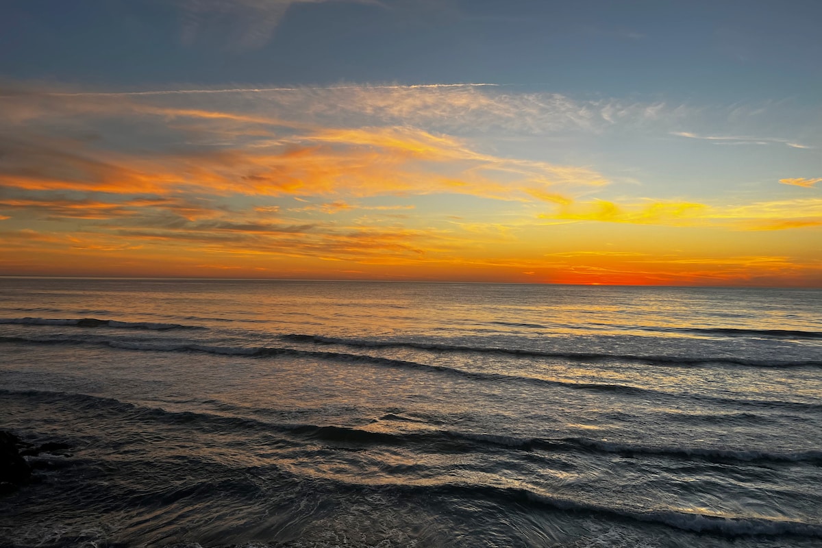 A serene ocean view is captured during sunset, with soft waves gently rolling onto the shore. The sky is adorned with hues of orange, pink, and blue, reflecting on the calm water's surface. The tranquil scene emphasizes the natural beauty of the beachfront.