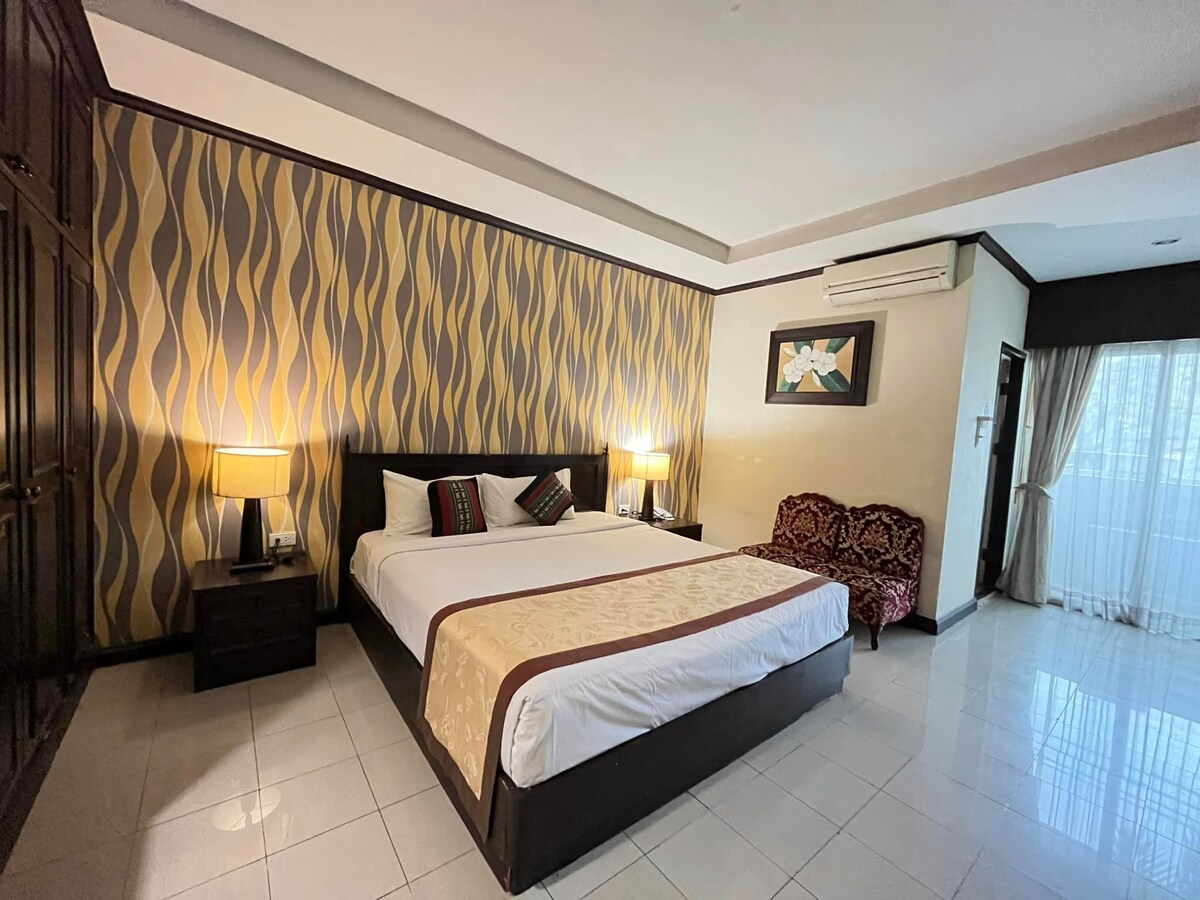 A spacious room features a large bed with neutral bedding, complemented by accent pillows. Two bedside lamps provide soft lighting. A vintage-style sofa is positioned against the wall, and the room is adorned with a patterned wallpaper, enhancing the overall design. Natural light filters through a large window.