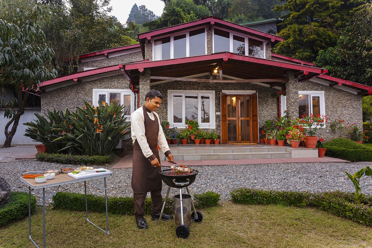 A charming villa is depicted in the image, surrounded by lush greenery. The outdoor area features a barbecue grill set up on a small table, indicating an inviting space for outdoor cooking. The front entrance showcases large windows and vibrant plant arrangements along the path.